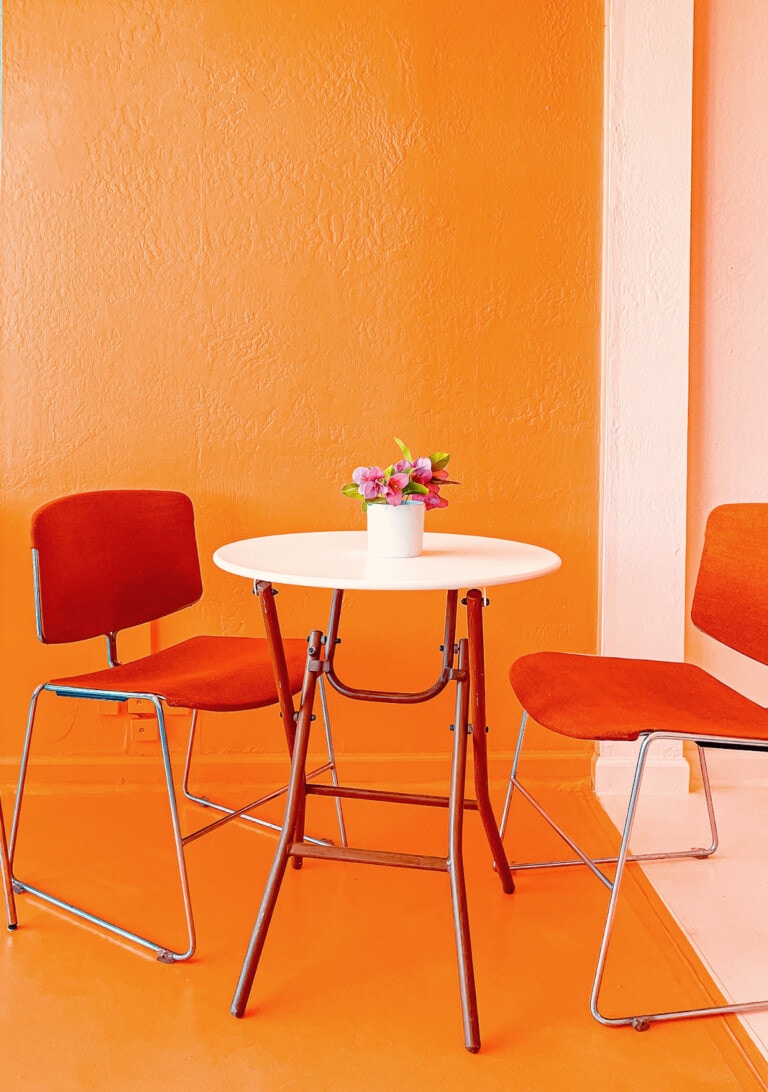 Rincón con pared y suelo naranja vibrante. Sillas rojas y mesa plegable blanca